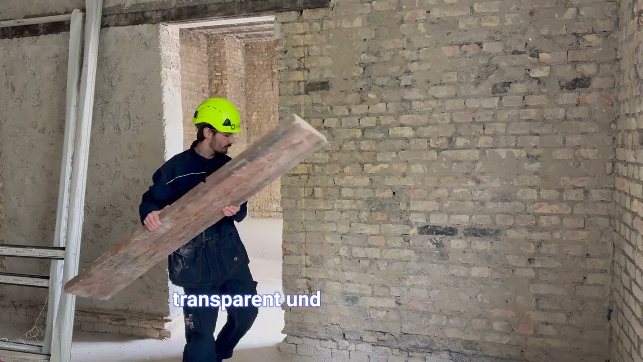 Standbild aus dem Brand-Video: Bauarbeiter mit Helm trägt eine Holzbohle durch einen Rohbau